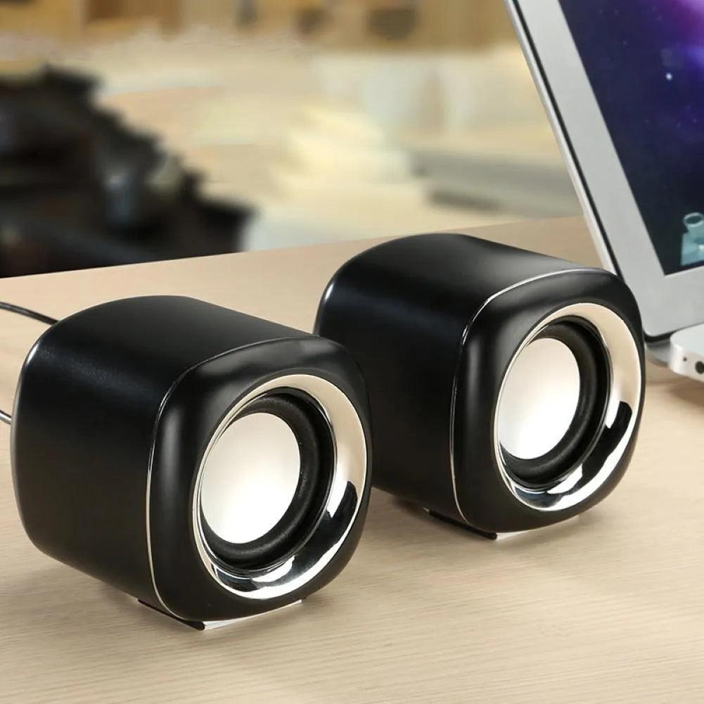A pair of compact black or white wired desktop computer speakers with a 3.5mm plug, shown next to a laptop.