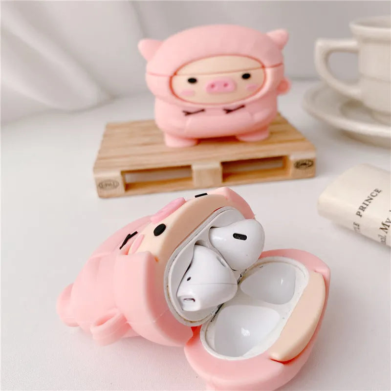 Pink silicone AirPods case with cartoon pig in sweater design, shown with flip lid open and a matching metal hook.