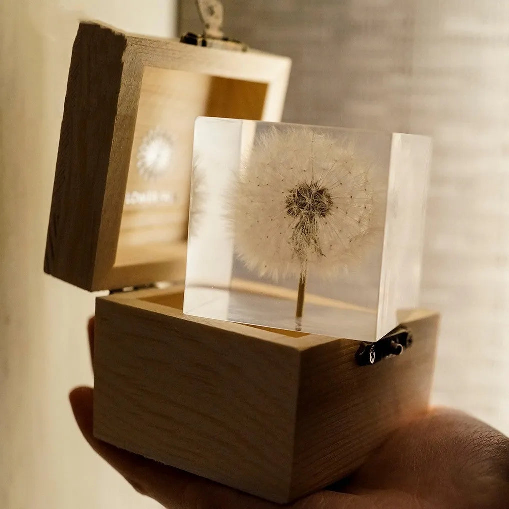 Clear cubic resin block with a dandelion flower inside, on a wooden box next to a USB charging base.