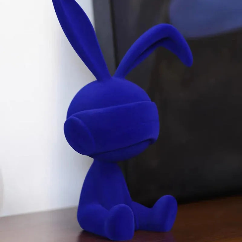 A resin table ornament of a rabbit wearing a suede blindfold, shown in blue, green, and red colour options.